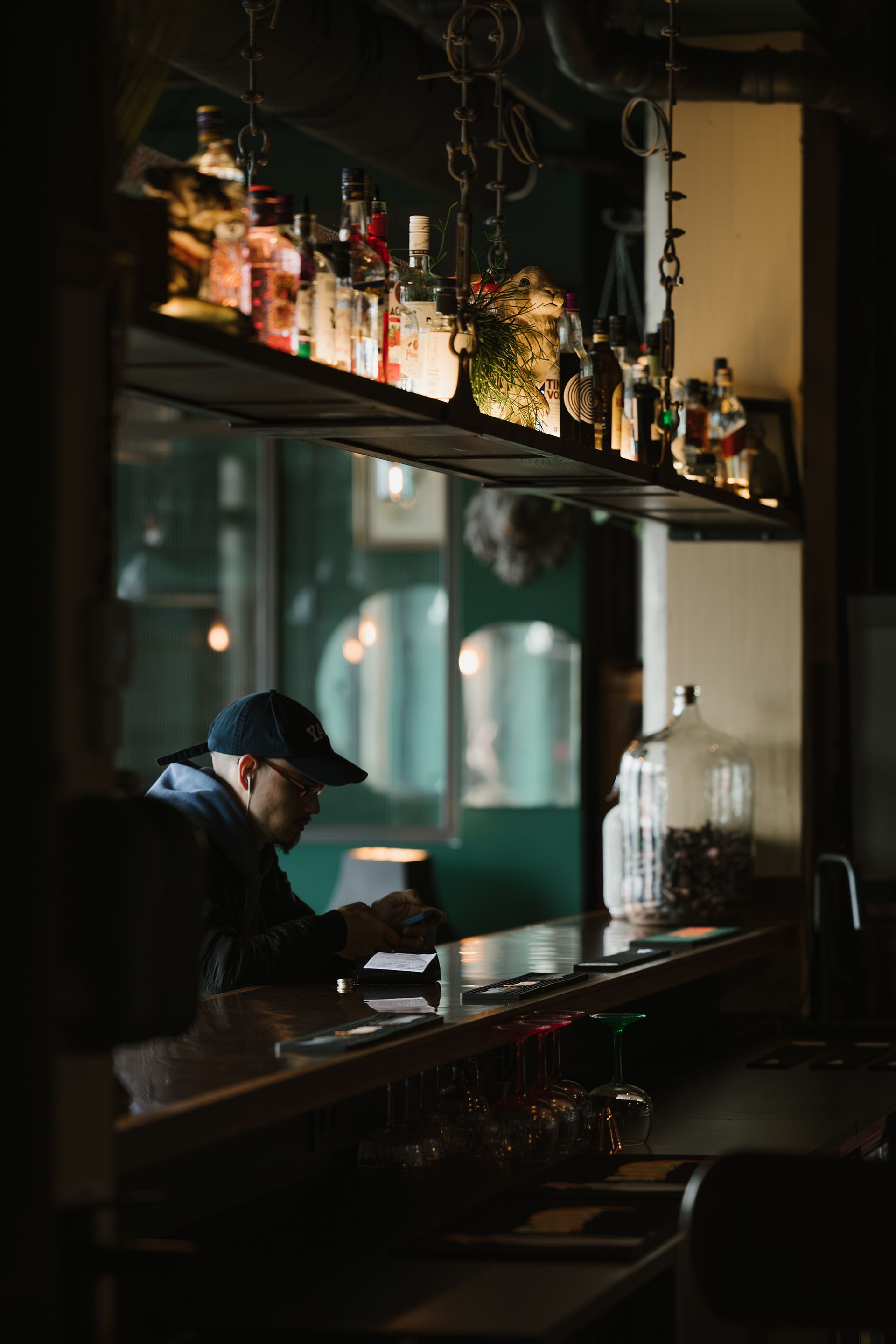 Person at a dimly lit bar, looking down. Bottles line the shelf. Dark wood and green tones.