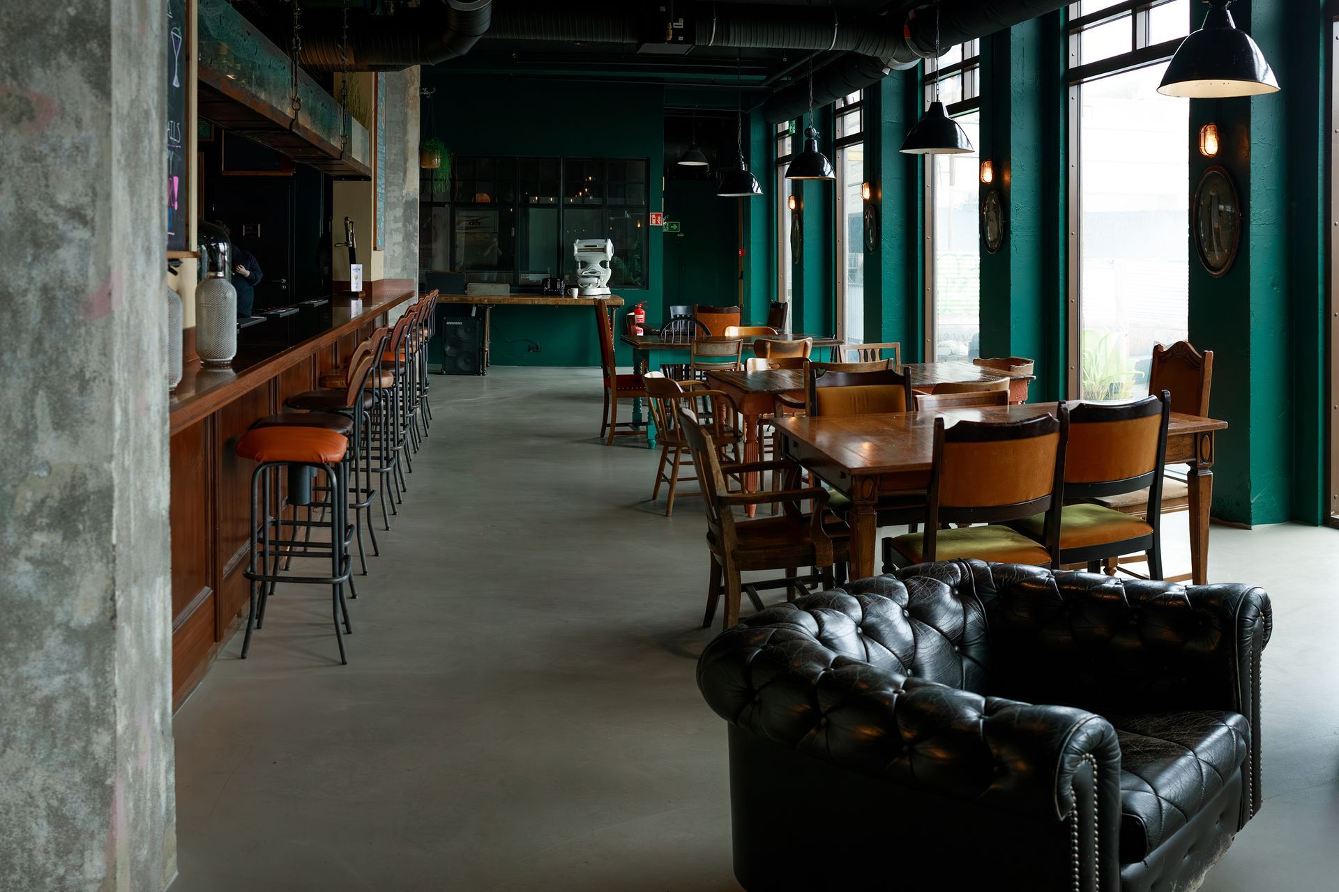 Interior of a bar/restaurant with a dark leather sofa, wooden tables, and a long bar with stools.