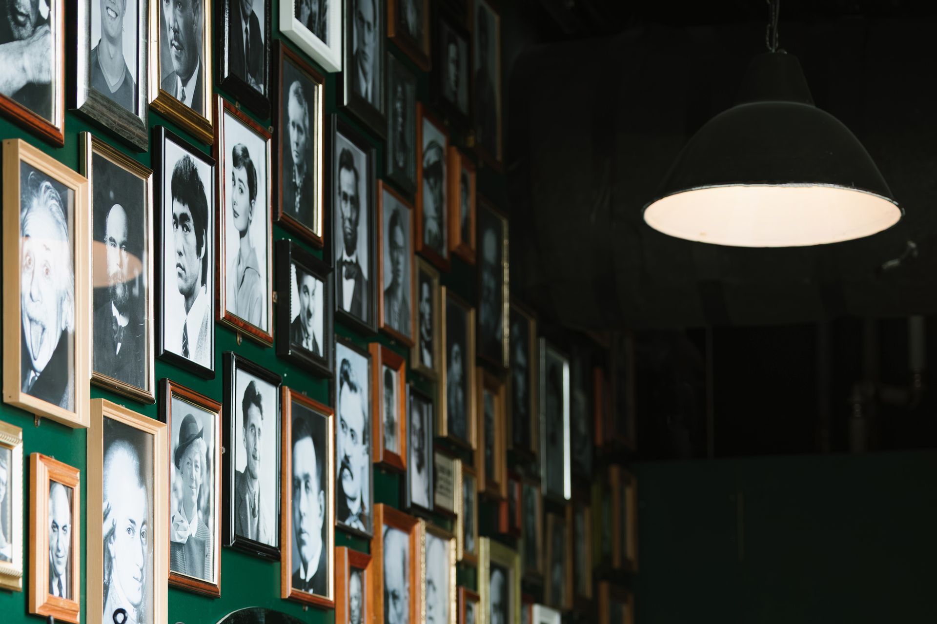Wall of framed black and white portraits under a hanging lamp in a dimly lit room.