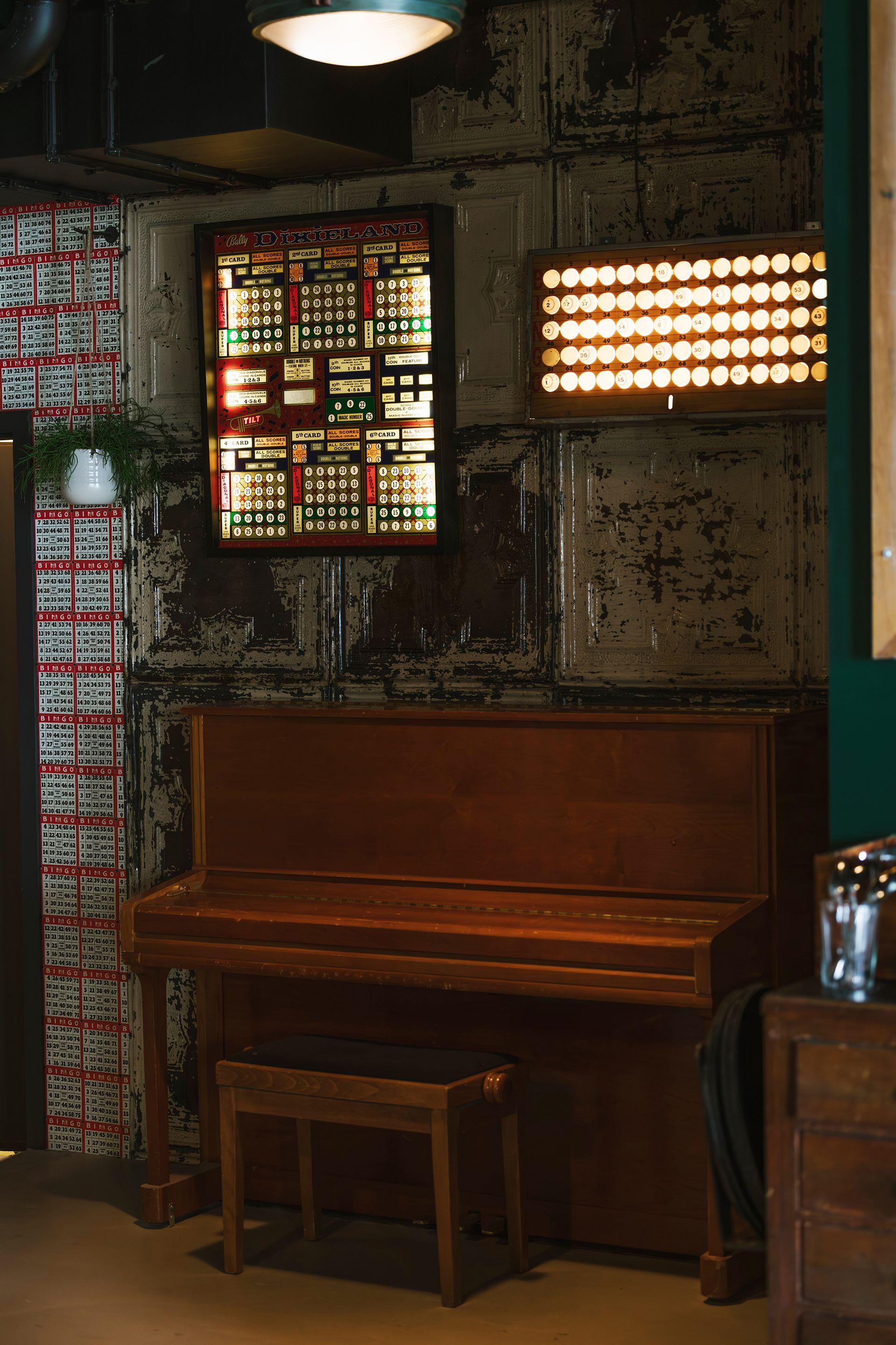 Piano in a rustic room with stained metal walls, illuminated by a hanging light and a window with colorful lights.