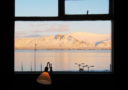 View of snowy mountains across a calm body of water, seen through a window. Lamp in foreground.