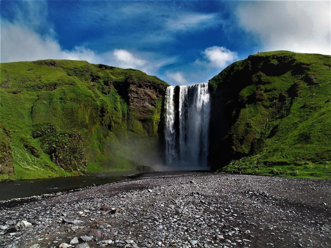 Waterfall cascading from a green, grassy cliff into a rocky foreground, with a blue sky.