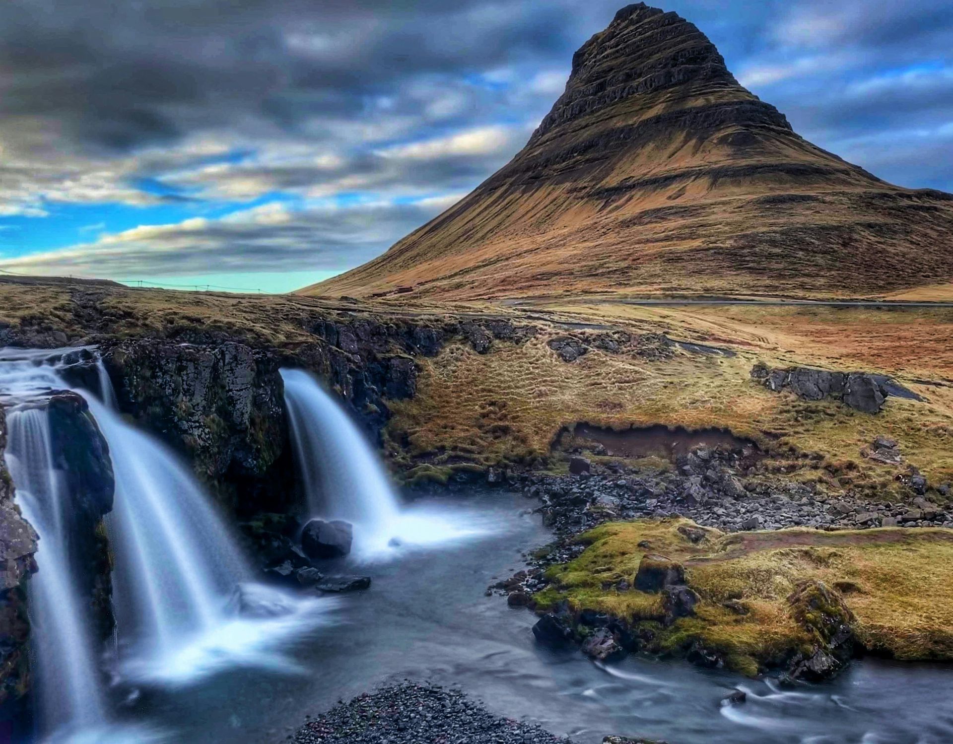 Waterfall cascading into a river with a large, cone-shaped mountain in the background under a cloudy sky.