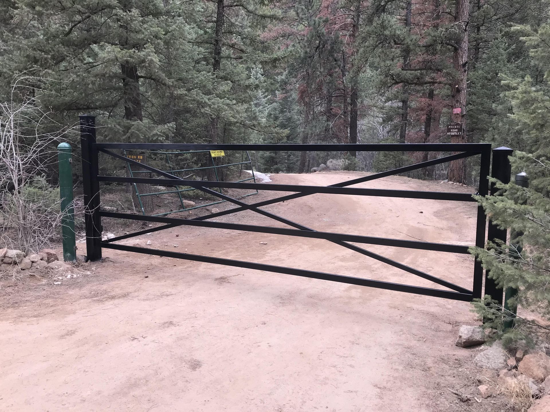 A black gate is closed on a dirt road in the woods.