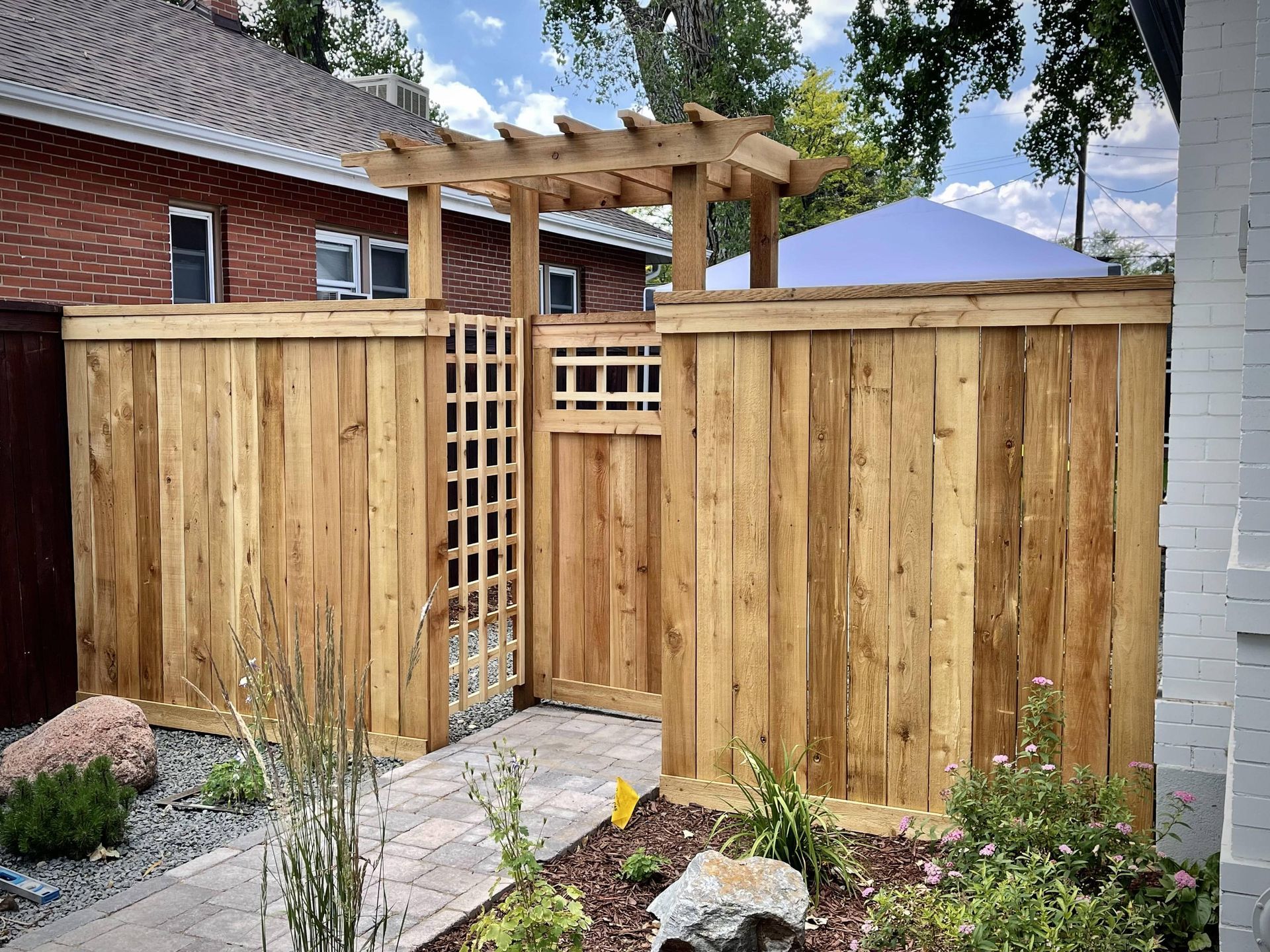 A wooden fence with a gate and a pergola in front of a brick house.