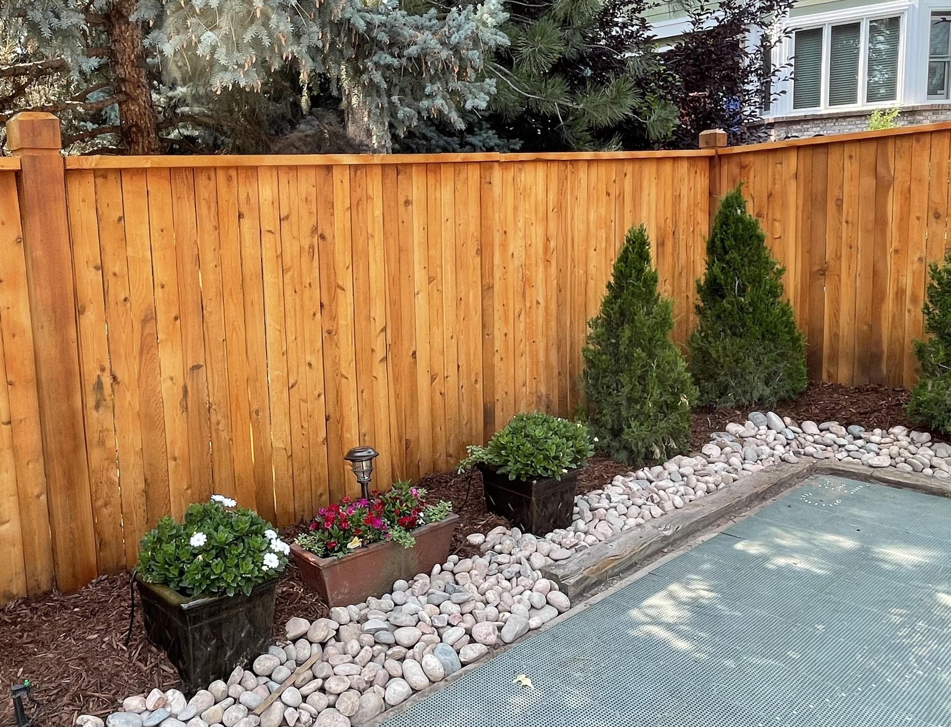 A wooden fence surrounds a swimming pool surrounded by rocks and flowers.