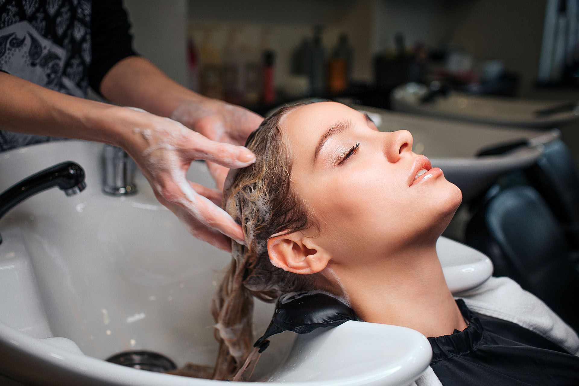 Woman getting hair washed at a salon. Hands lather shampoo into her hair in a white sink, eyes closed.