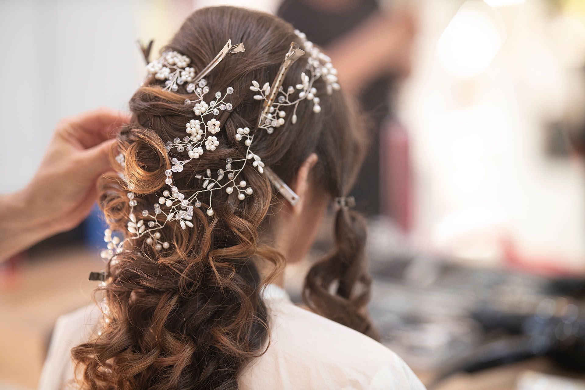 A woman has her hair styled with ornate floral hairpins. A stylist is adjusting the hair.