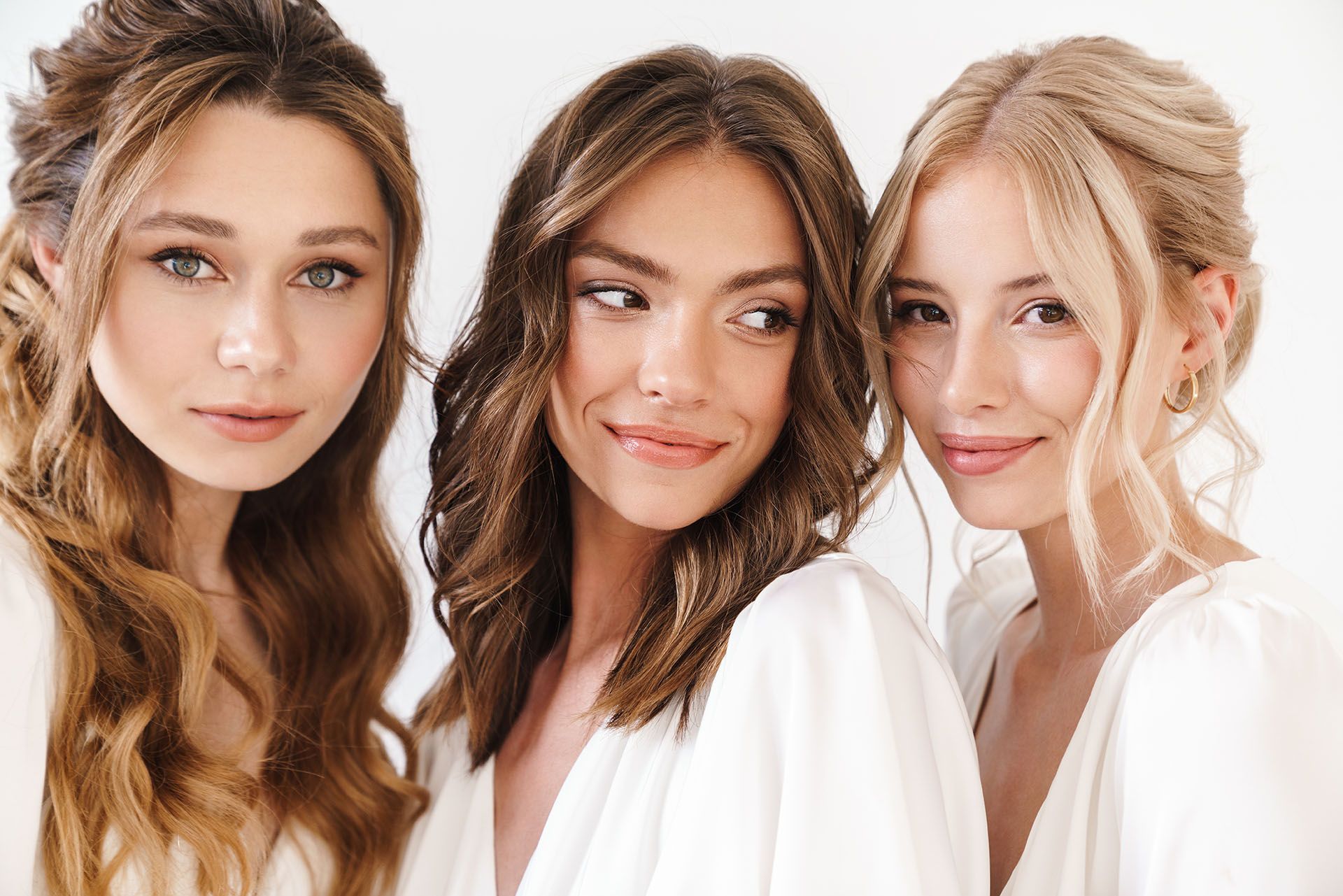 Three women in white, smiling, looking at the camera. Two have up-dos, one has loose waves.