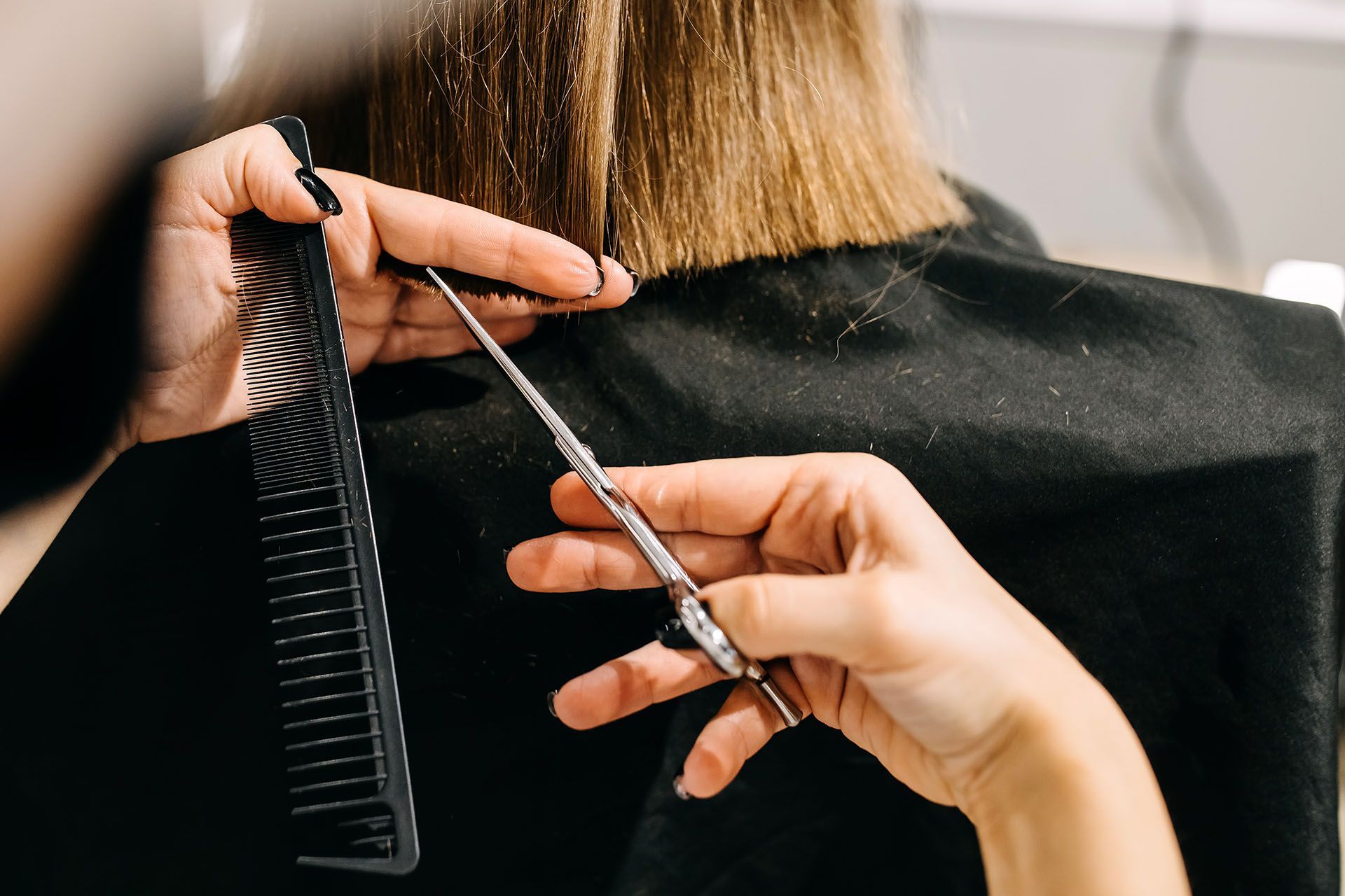 Hairdresser cutting client's straight blonde hair with scissors, using a comb. Black cape, indoor setting.