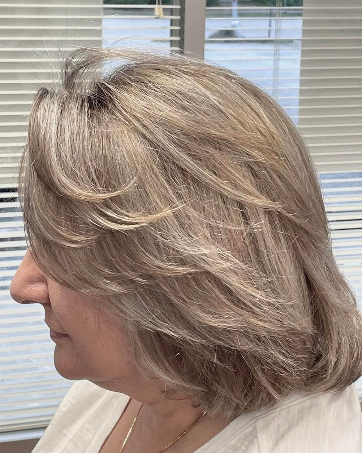Woman with layered, ash blonde hair, profile view. Indoor setting with window blinds.