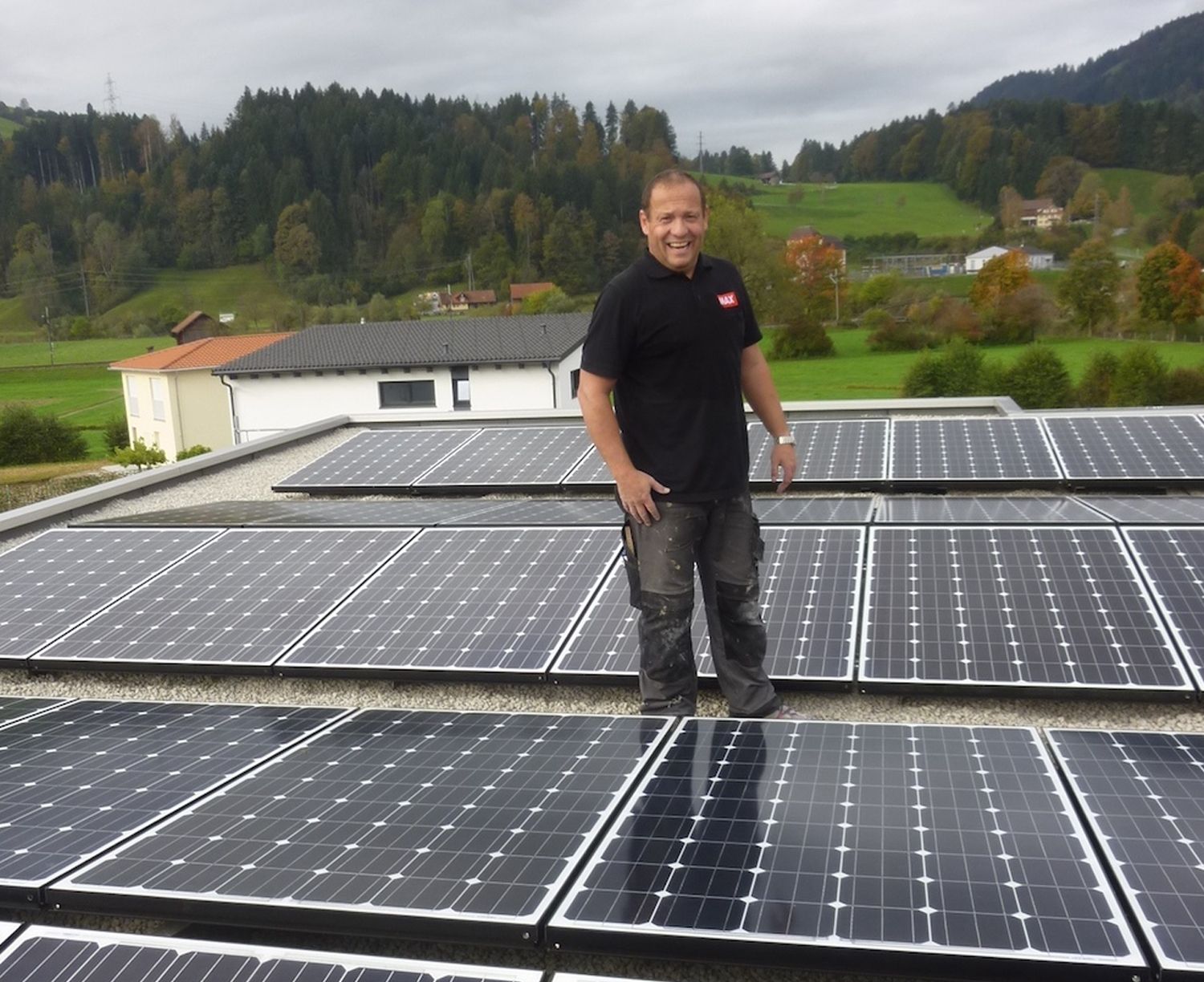 Lächelnder Mann steht auf einem mit Solarmodulen bedeckten Dach, im Hintergrund ein grüner Hügel.