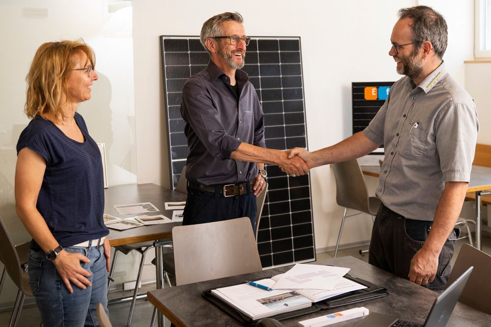 Drei Personen in einem Raum mit Solarmodulen. Zwei Männer schütteln sich die Hände, eine Frau schaut zu.