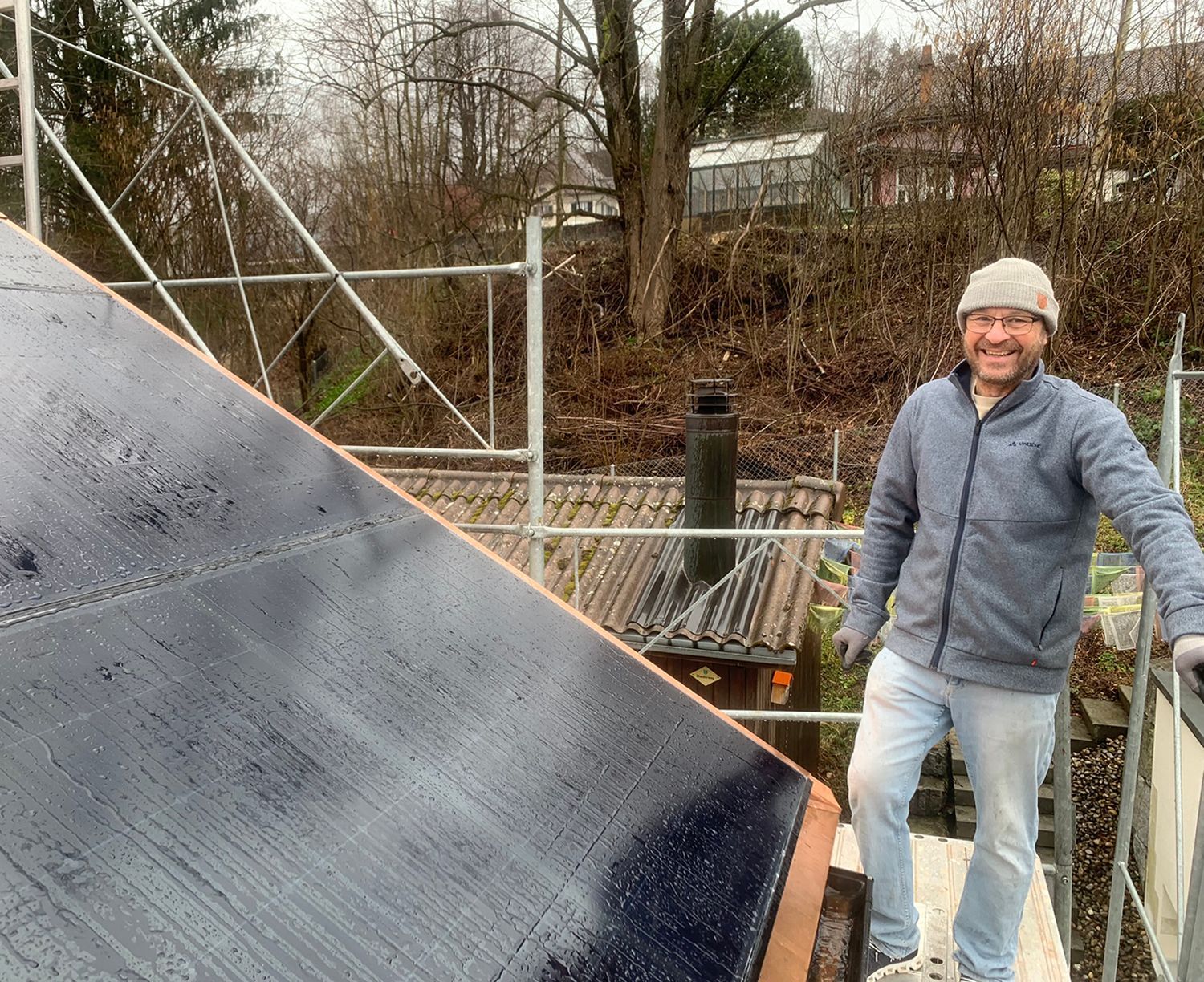 Mann auf einem Dach mit Solarpanel. Er trägt Jacke, Jeans und Mütze und lächelt. Im Hintergrund ein Gerüst.