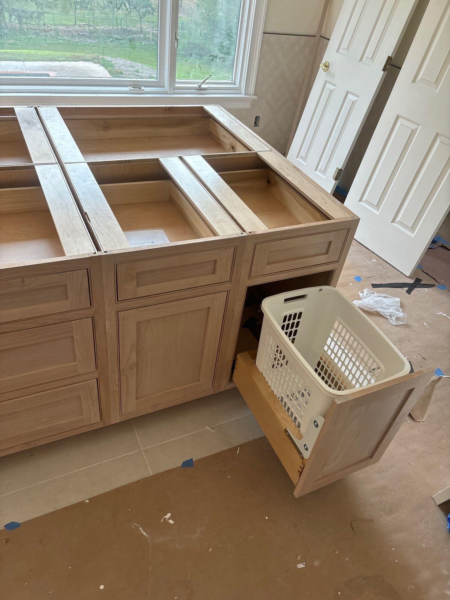 A partially-constructed kitchen island with cabinets and a pull-out laundry basket; light wood, indoors.