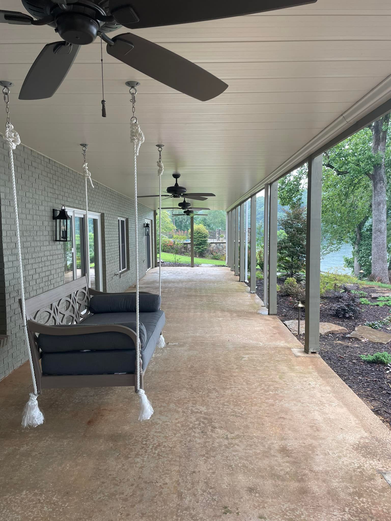 Covered porch with hanging swings, gray brick wall, and view of trees.