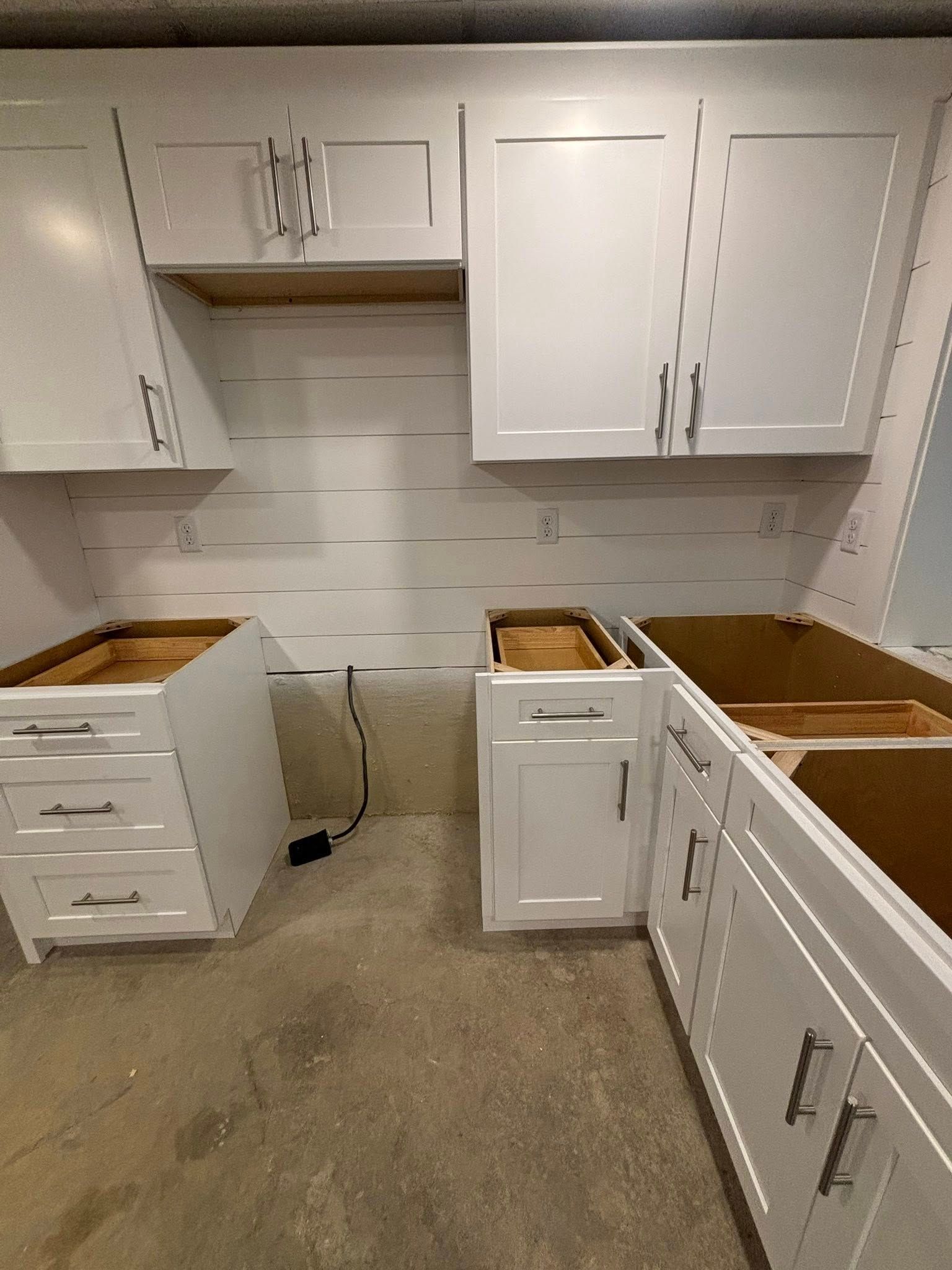 White kitchen cabinets being installed; white shiplap backsplash, concrete floor.