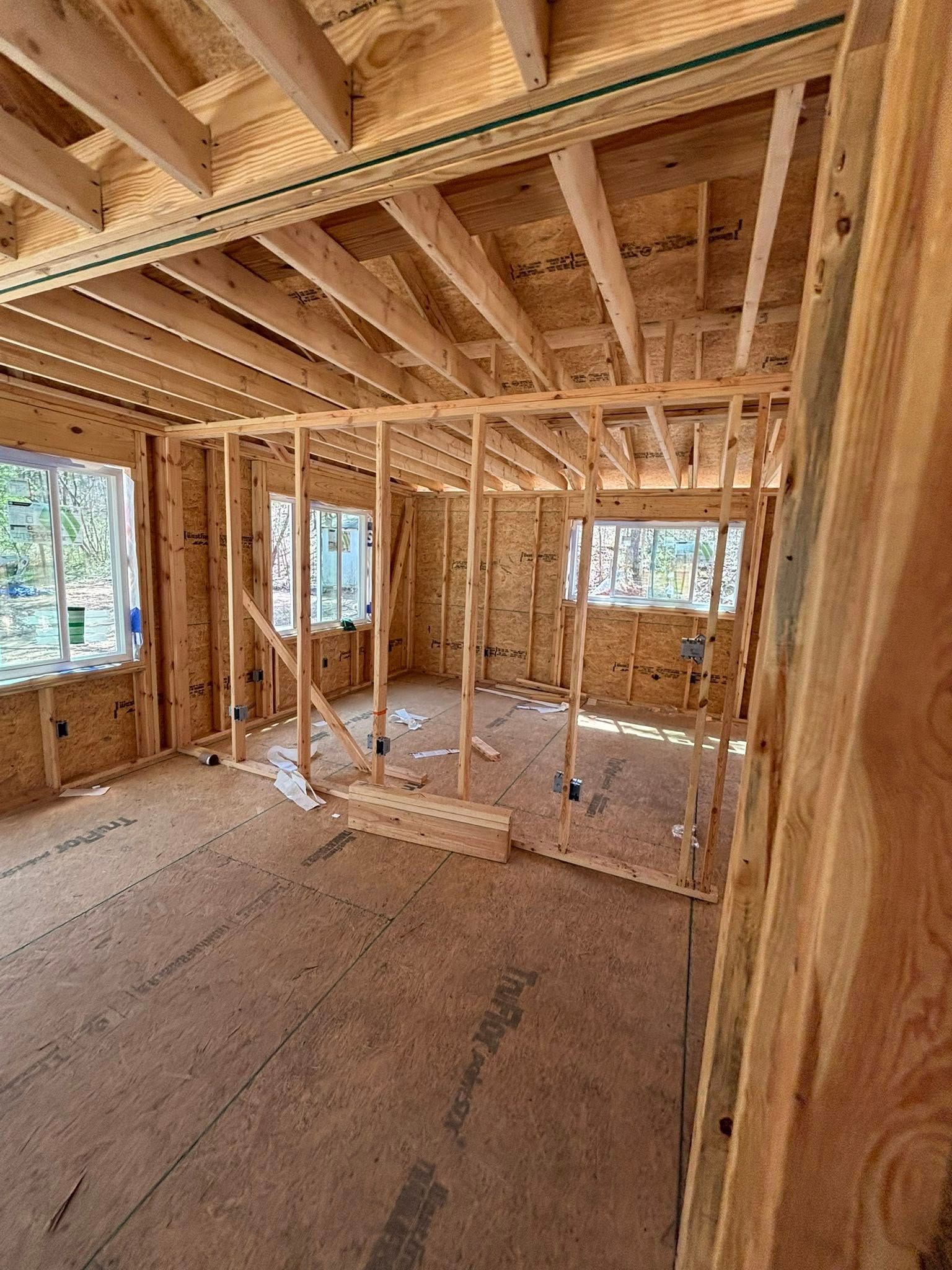 Interior view of a wooden house under construction. Studs, rafters, windows, and OSB flooring are visible.