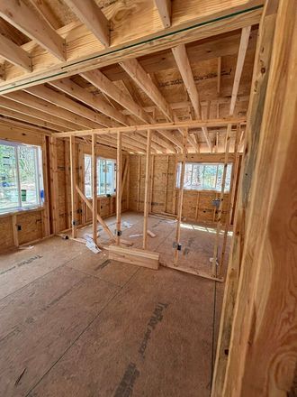 Interior view of a wooden house under construction. Studs, rafters, windows, and OSB flooring are visible.