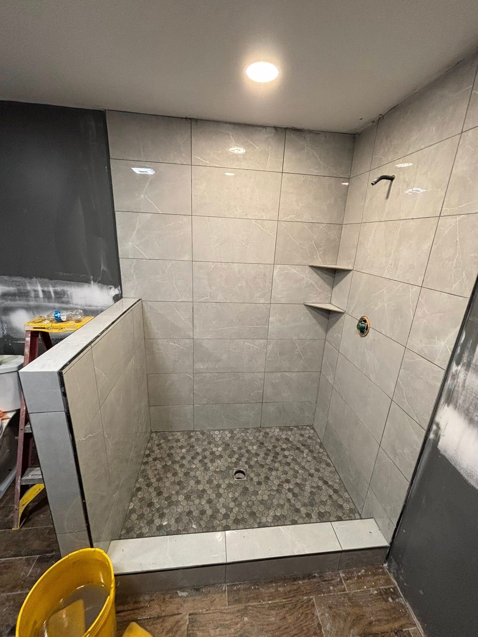 Shower stall under construction, gray tiled walls, pebble floor, built-in shelves.