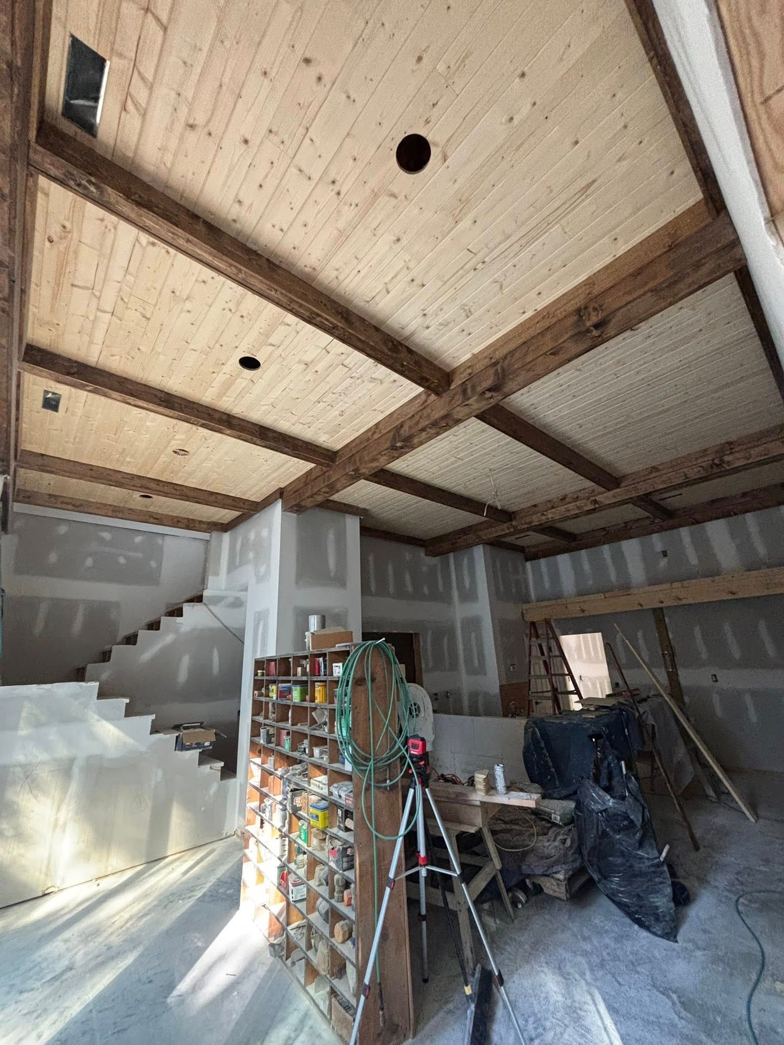 Construction interior with exposed wooden beams and ceiling, unfinished drywall, ladder, and a shelf of paint cans.