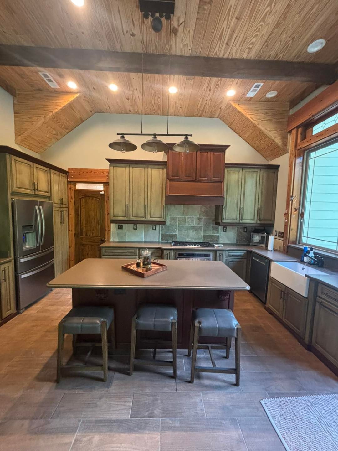 Rustic kitchen with wood beams, green cabinets, island, stools, and stainless steel appliances.