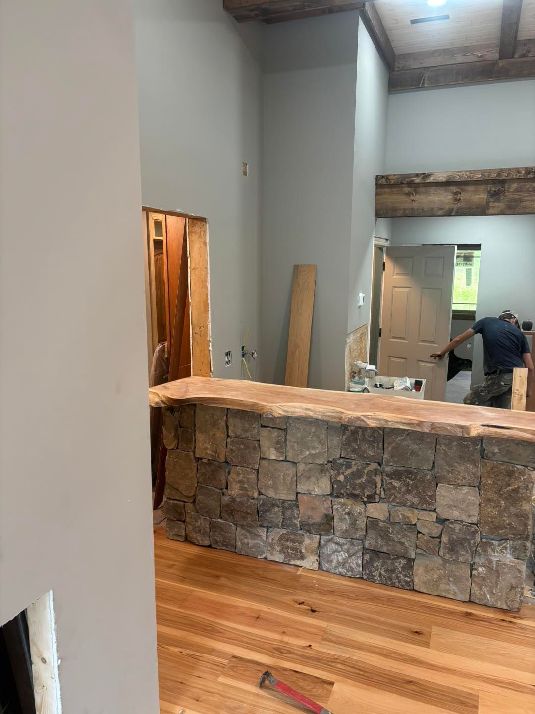 Interior under construction: stone-faced bar with wood countertop, wooden beams, light wood floor, gray walls.