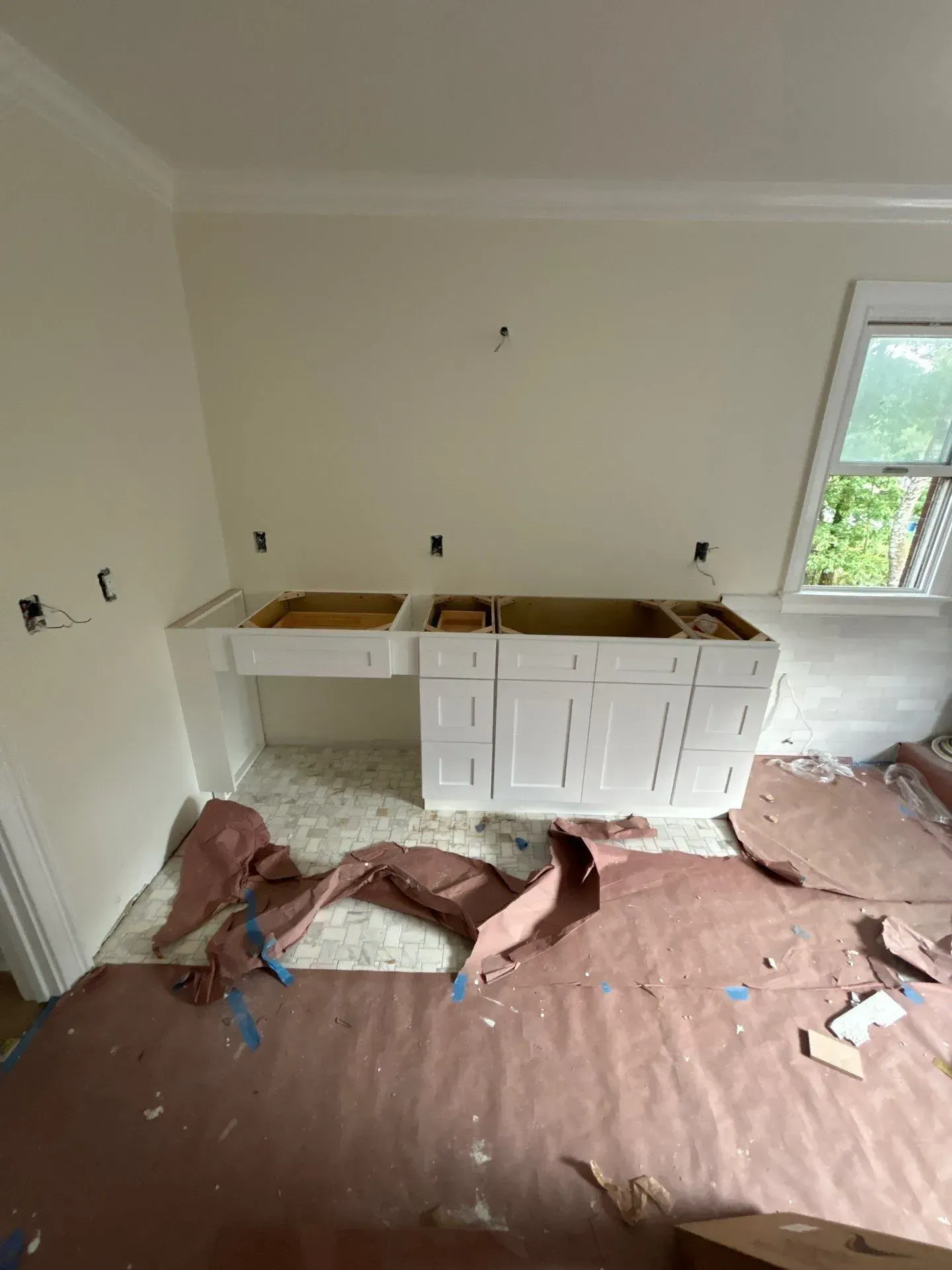 Bathroom renovation in progress. White cabinets, exposed electrical outlets, brown flooring protection, window.