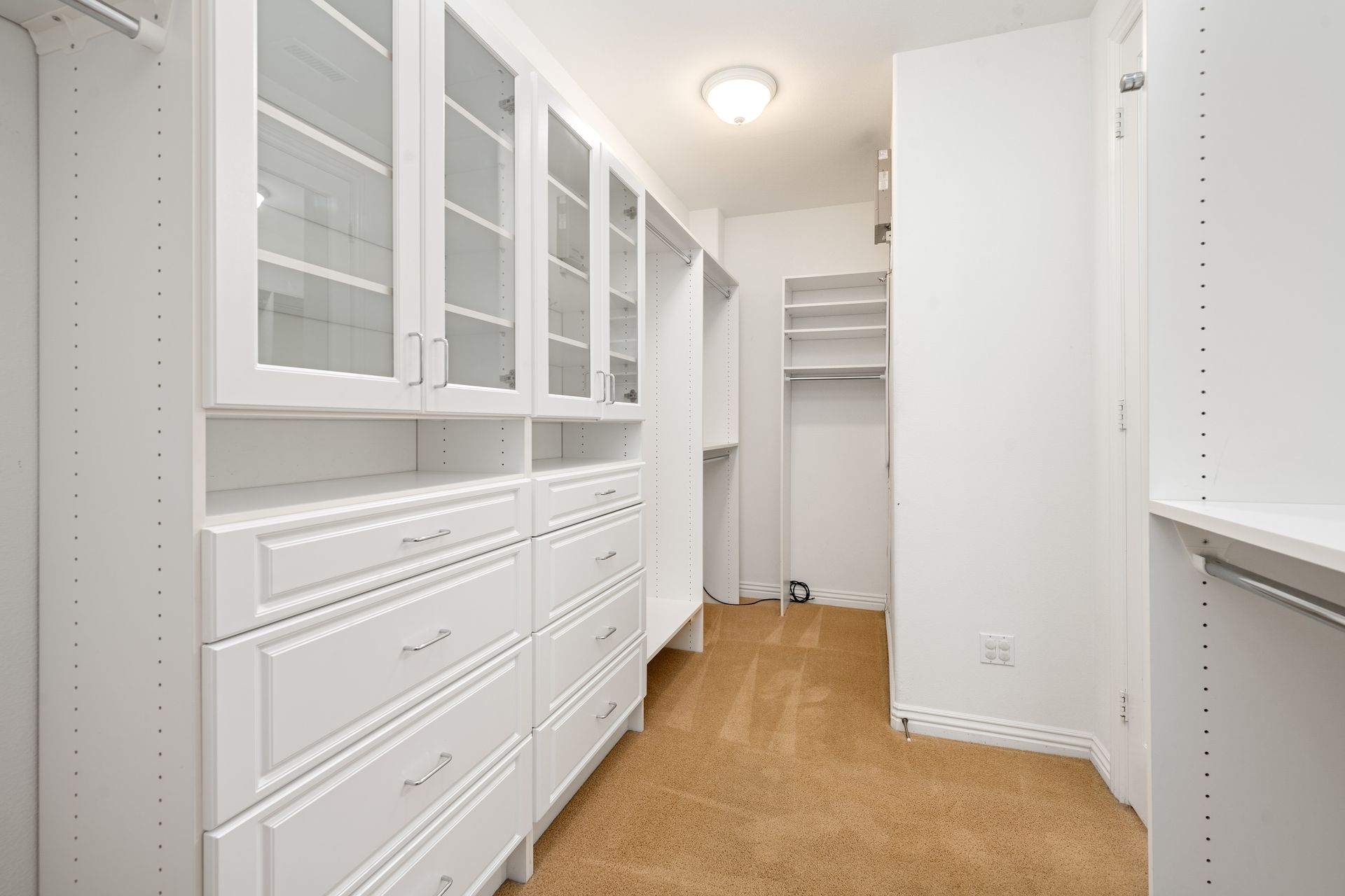 White walk-in closet with drawers and glass-door cabinets; tan carpet, bright overhead lighting.