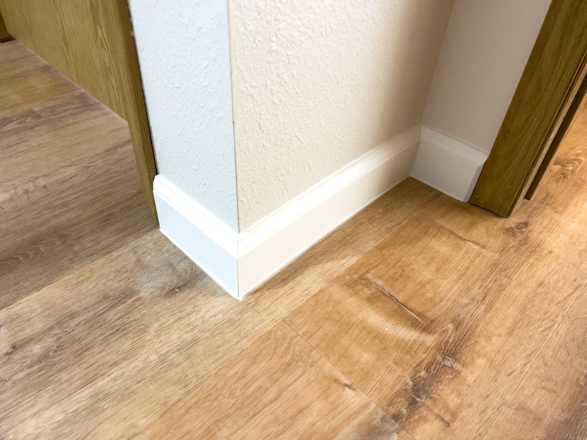 Corner of a room with light wood-look flooring, white baseboards, and textured white wall.
