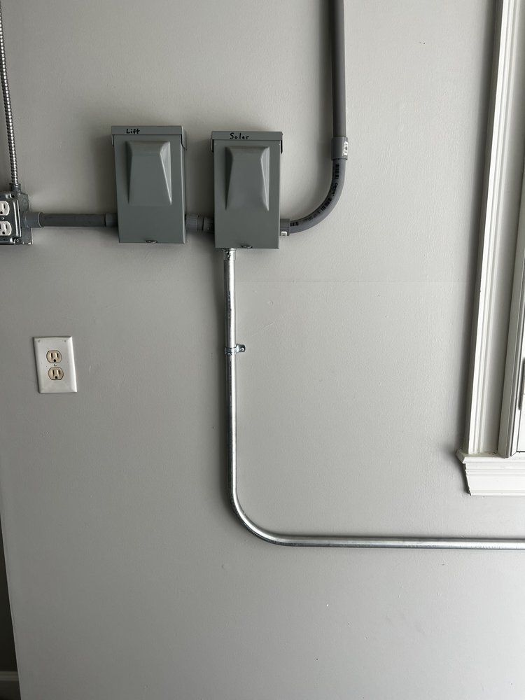 Gray electrical boxes and conduit mounted on a wall beside an outlet and a fluorescent light fixture