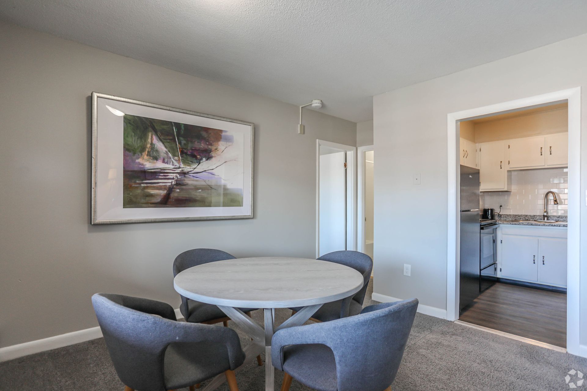 Dining area with a round table, four gray chairs, and a landscape painting. Kitchen is visible in the doorway.