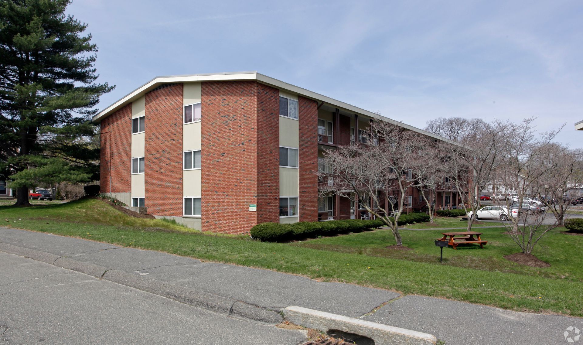 Three-story brick apartment building on a grassy hill; trees, sky, and road in view.