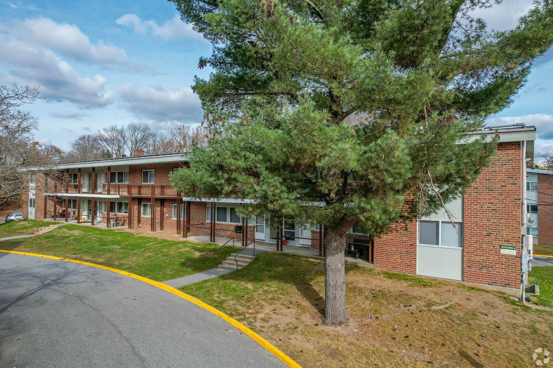 Apartment building exterior, brick and siding with a large tree in front, on a sloped grassy lot, overcast day.