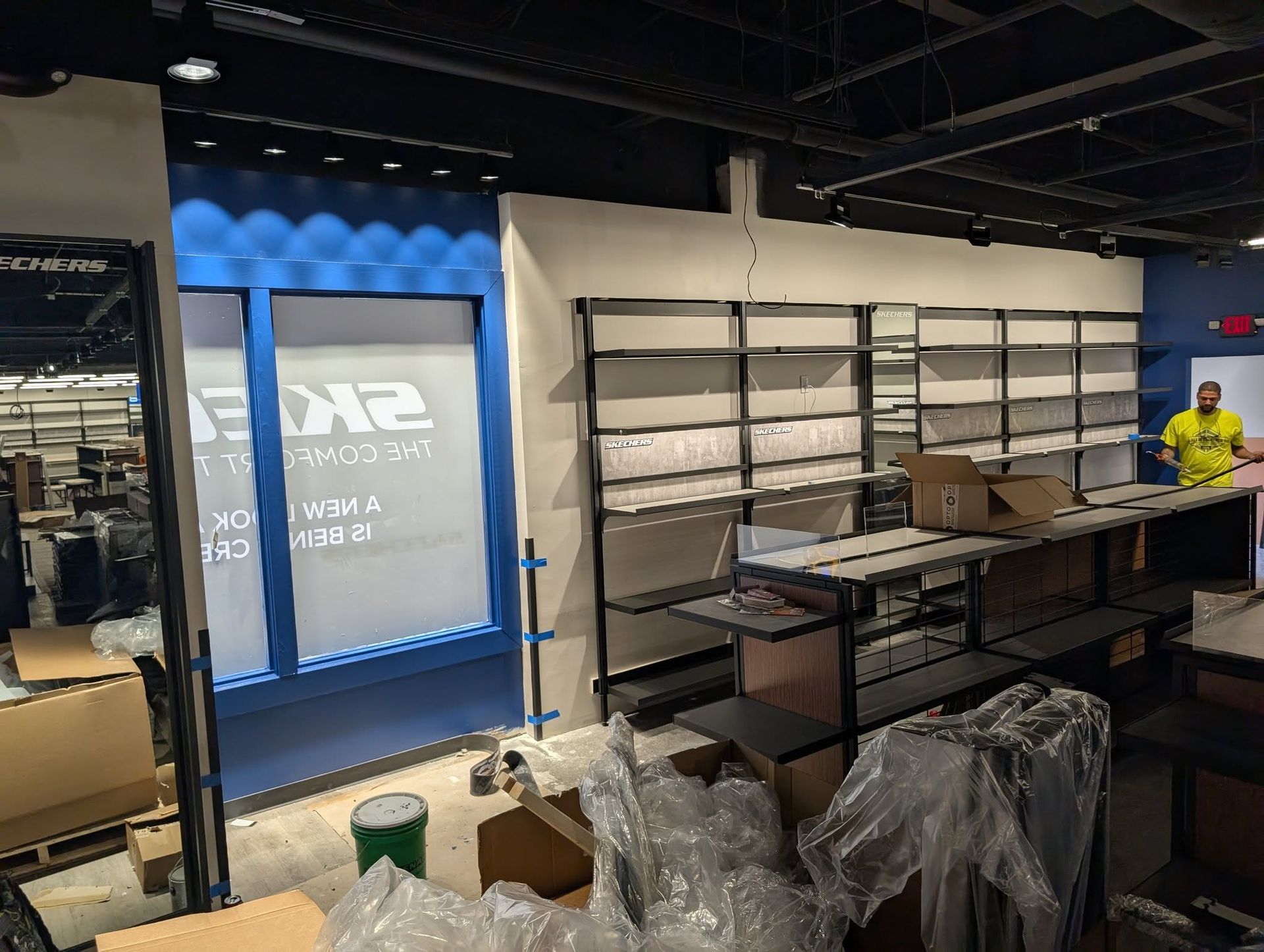 Interior of a store being set up; shelves and displays under construction; a worker in yellow shirt.