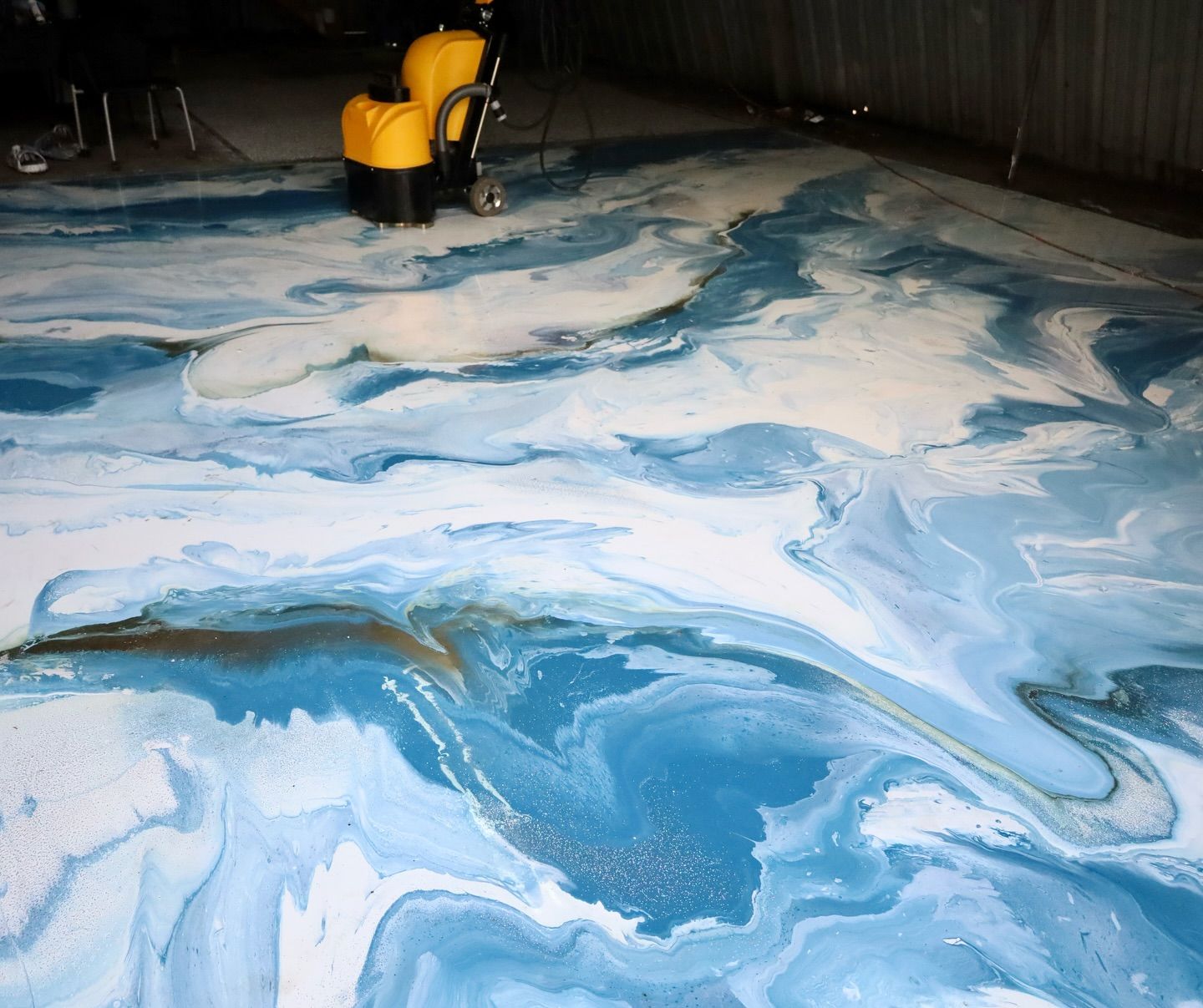 Blue, white, and gold epoxy floor with swirling patterns, resembling waves. A yellow machine is in the background.