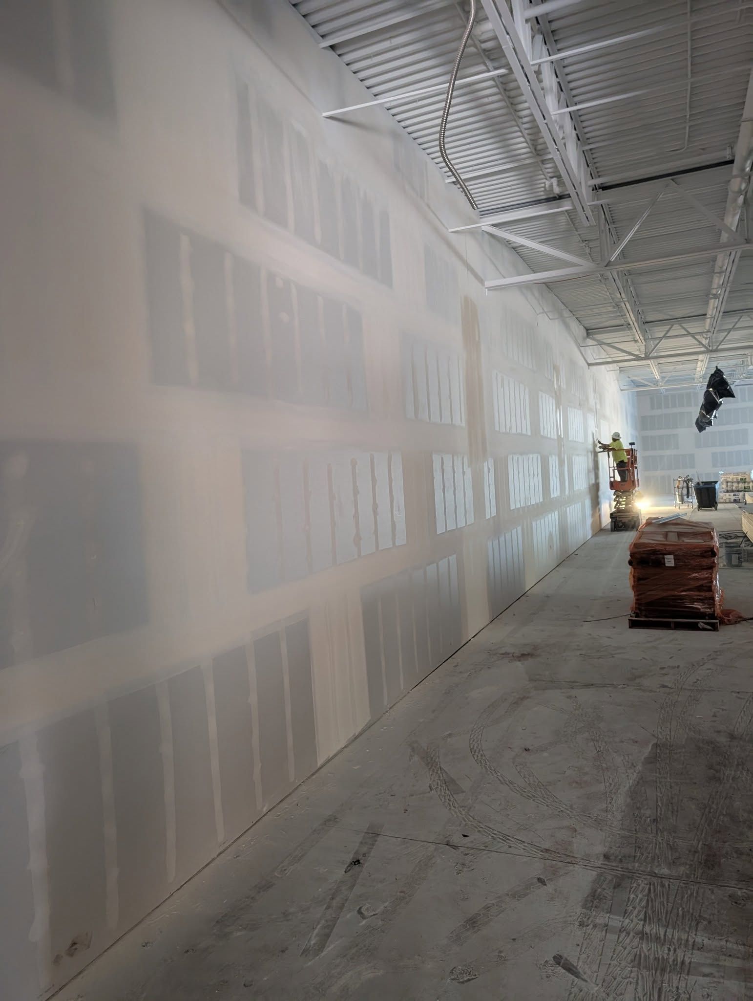 Interior construction site with drywall on wall. Concrete floor, exposed ceiling, and materials visible.
