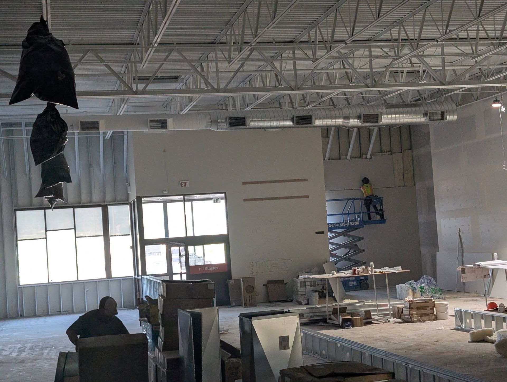 Construction site interior: Workers near drywall, windows, and metal scaffolding; exposed ceiling beams overhead.