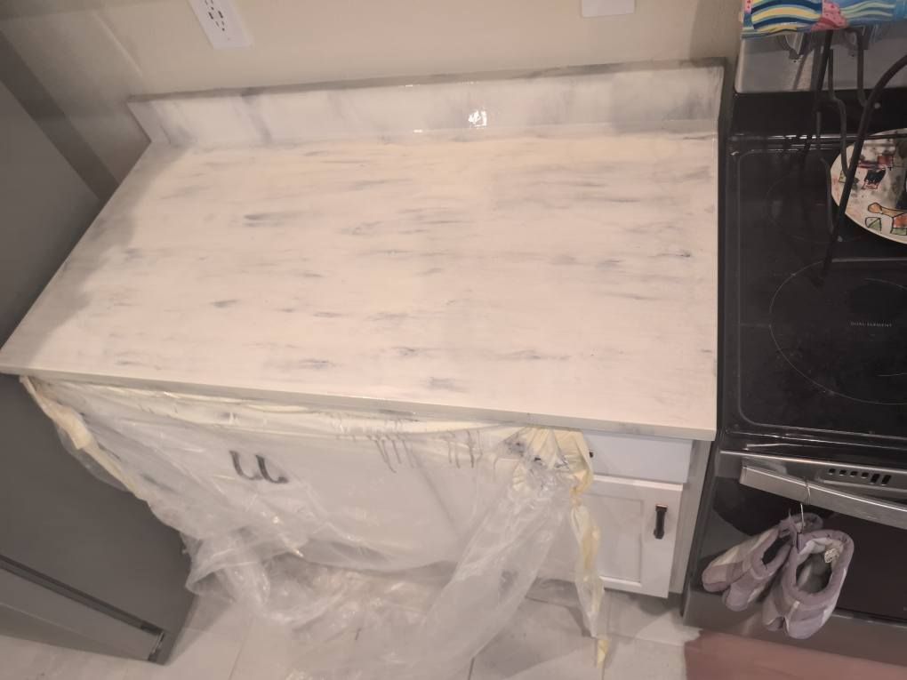 White kitchen countertop with a distressed paint finish, cabinets covered in plastic, next to a black oven.