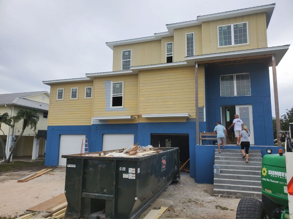 Two-story house under construction, yellow and blue exterior, people walking up stairs, a dumpster in the front.
