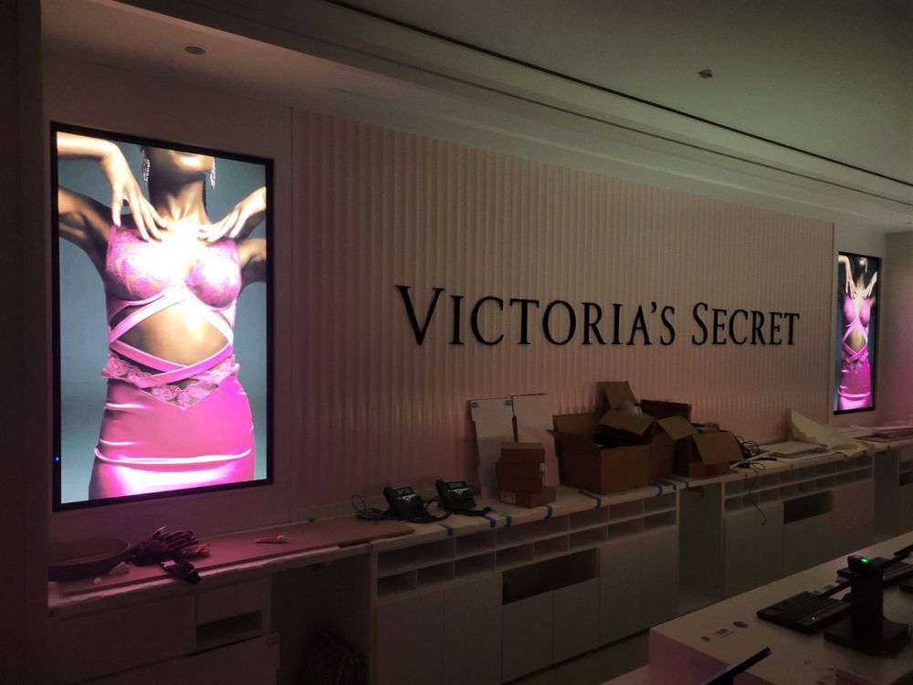 Victoria's Secret store interior. Large logo and pink lingerie ads on wall. Boxes and unfinished shelving visible.