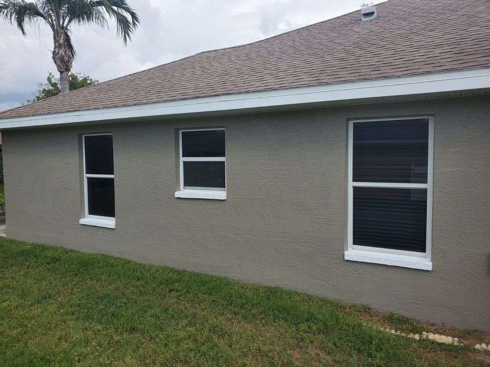 Exterior of a house with three windows, light brown stucco walls, white trim, and brown roof.