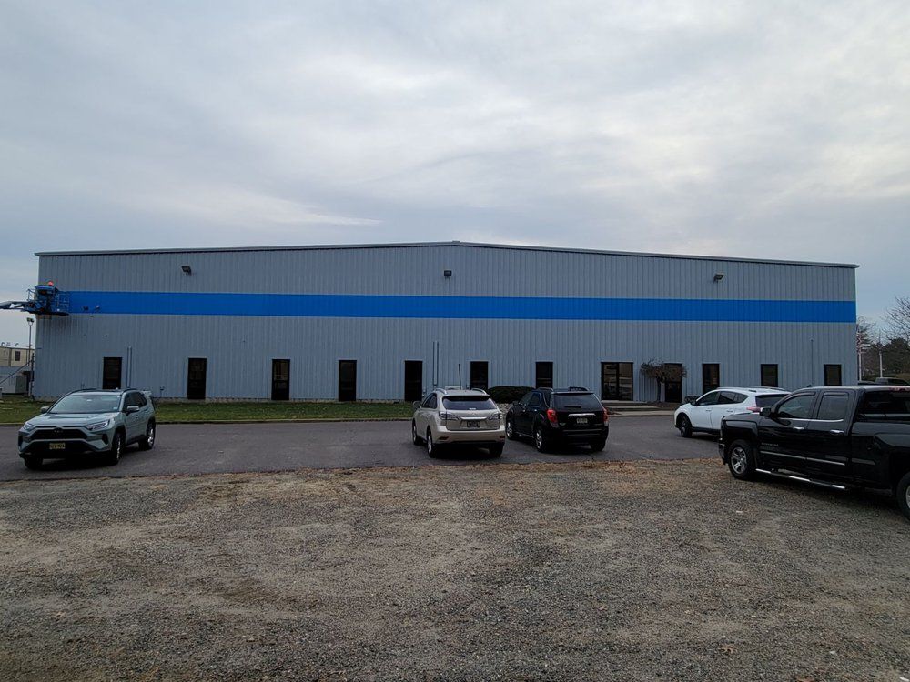Gray industrial building with blue stripe, parked cars in front, overcast sky.
