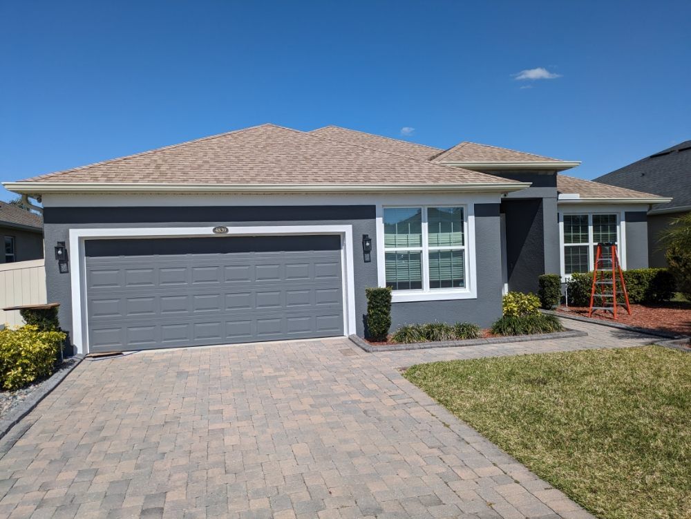 Gray house with a tan roof and gray garage door. Brick driveway and green lawn.