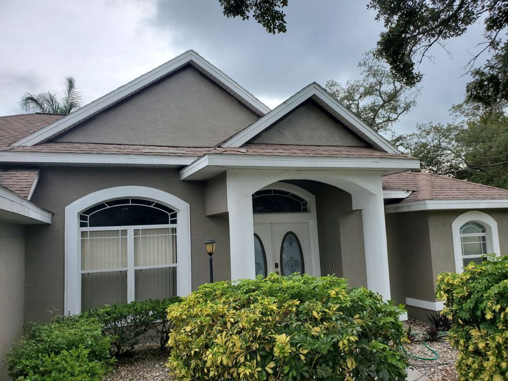 Beige house with white trim, a brown roof, and lush green bushes. Cloudy sky.
