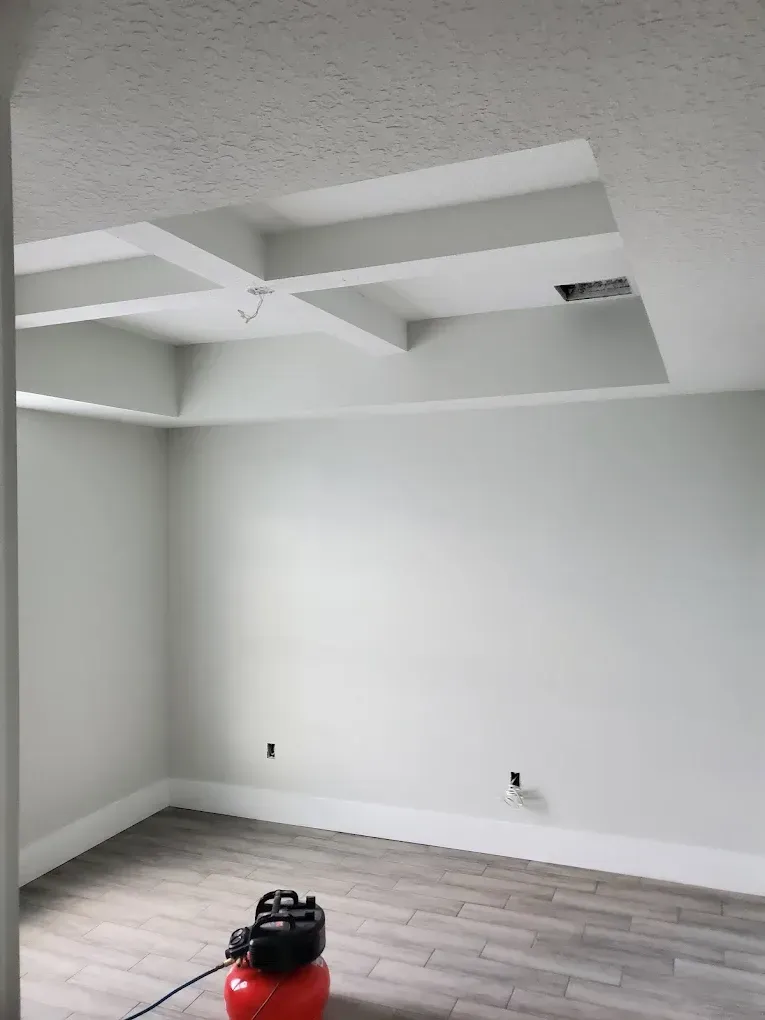 Empty room with gray walls, white ceiling with beams, and gray wood-look flooring; air compressor in foreground.