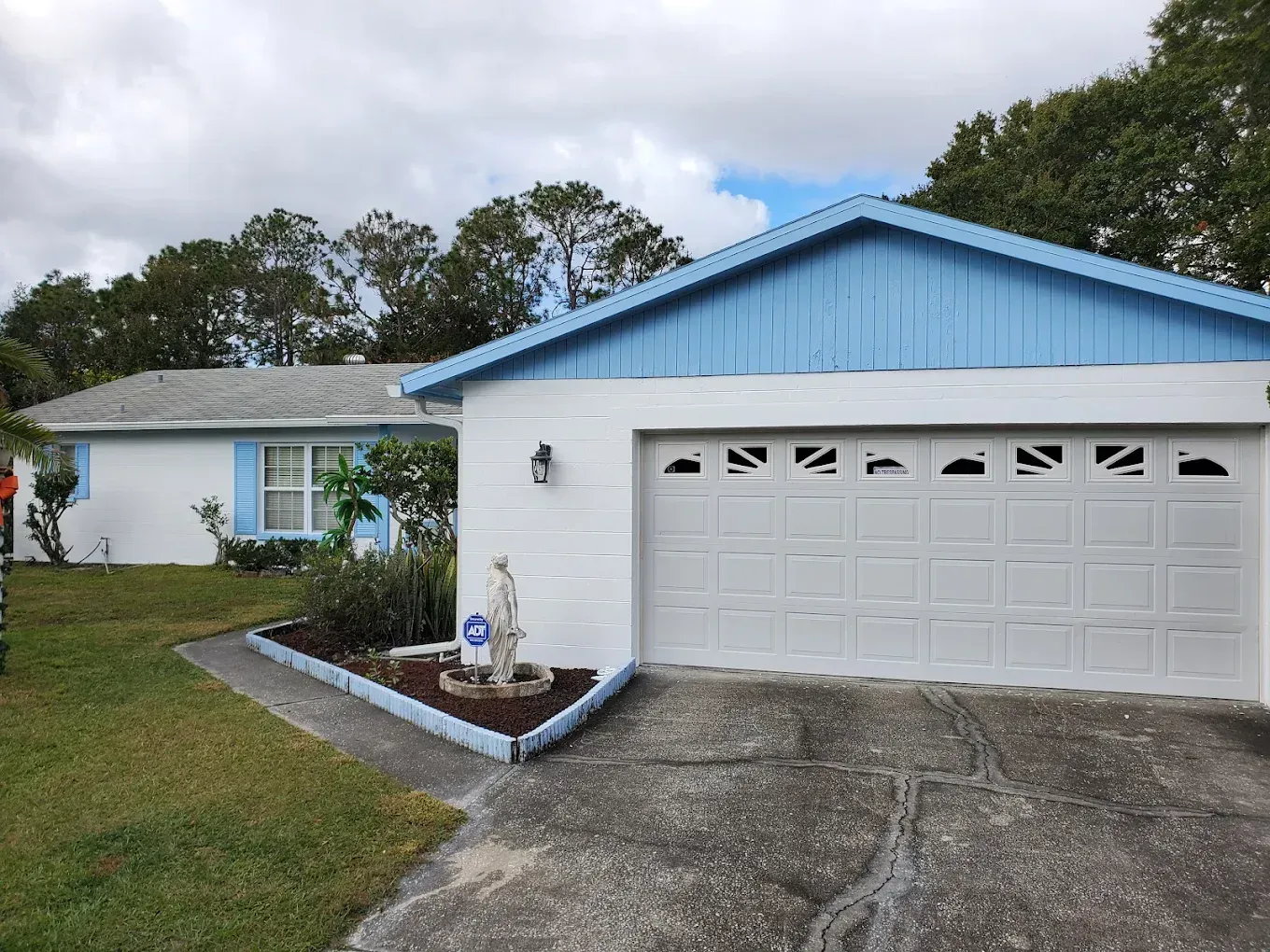 White house with blue trim, two-car garage, statue in garden bed. Cloudy day.