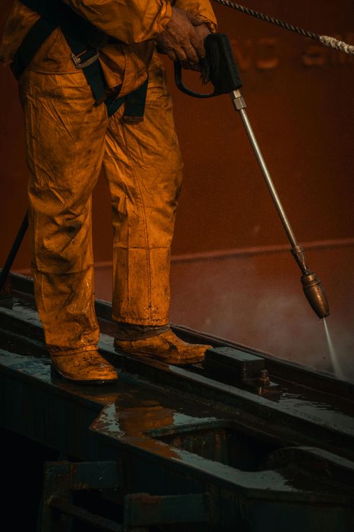 Worker in orange rain gear pressure-washing a dark metal surface at night