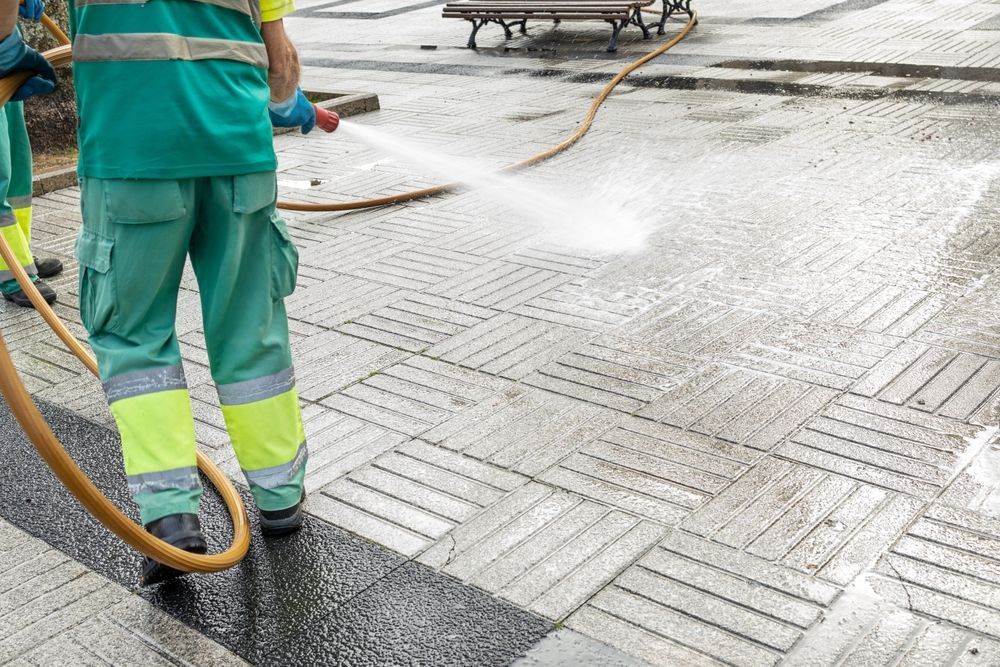 Worker in teal uniform pressure-washing a wet sidewalk with a hose