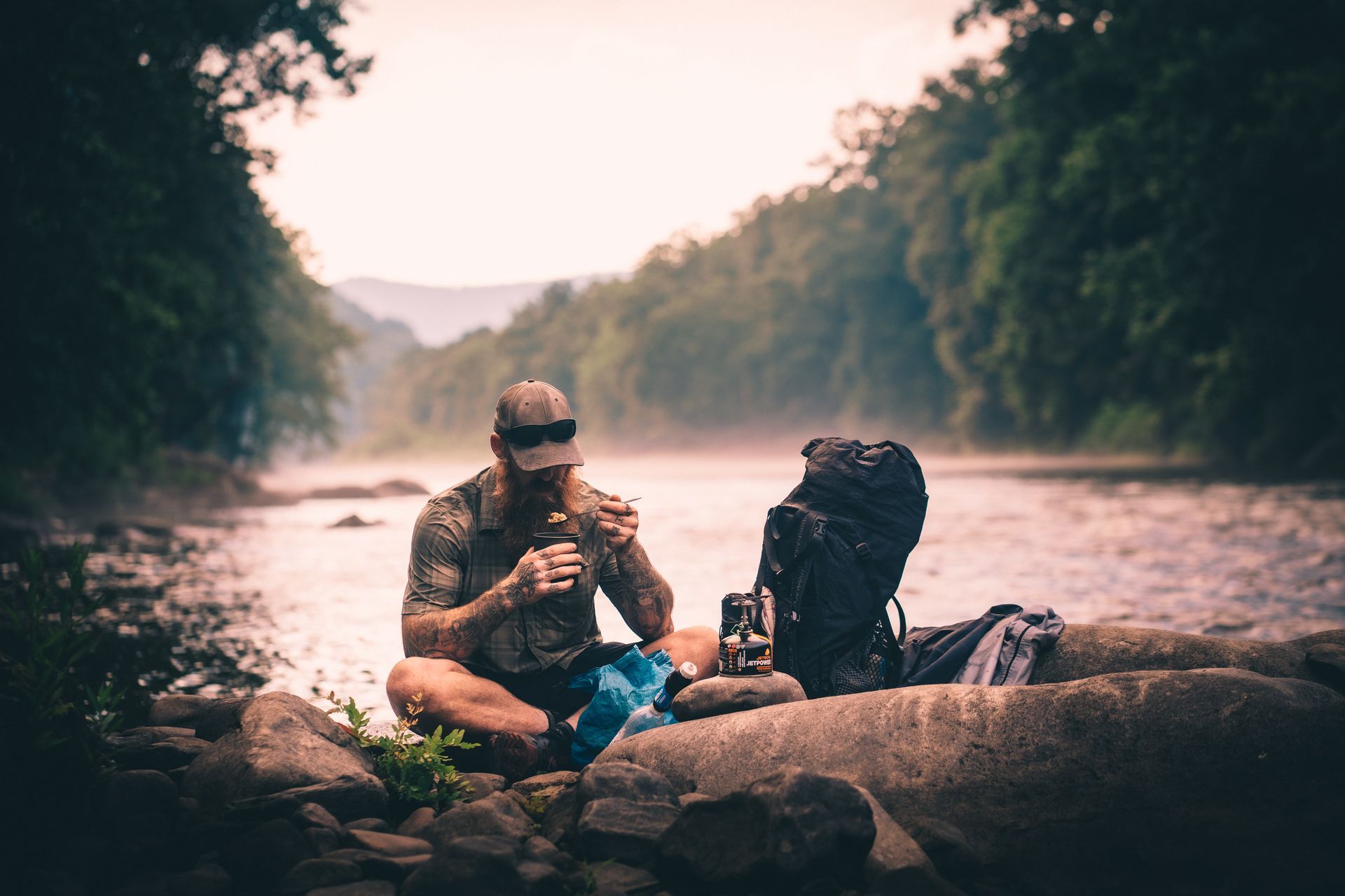A man is sitting on a rock by a river eating a sandwich.